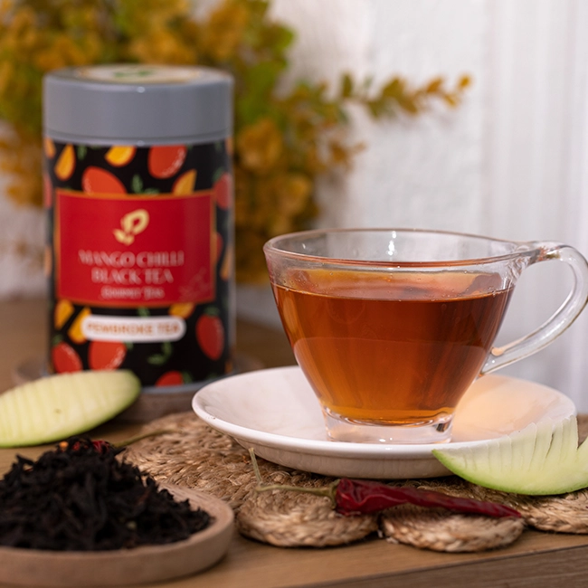 Mango chilli black tea in a glass cup with dried tea leaves, sliced mango, and tin packaging, India